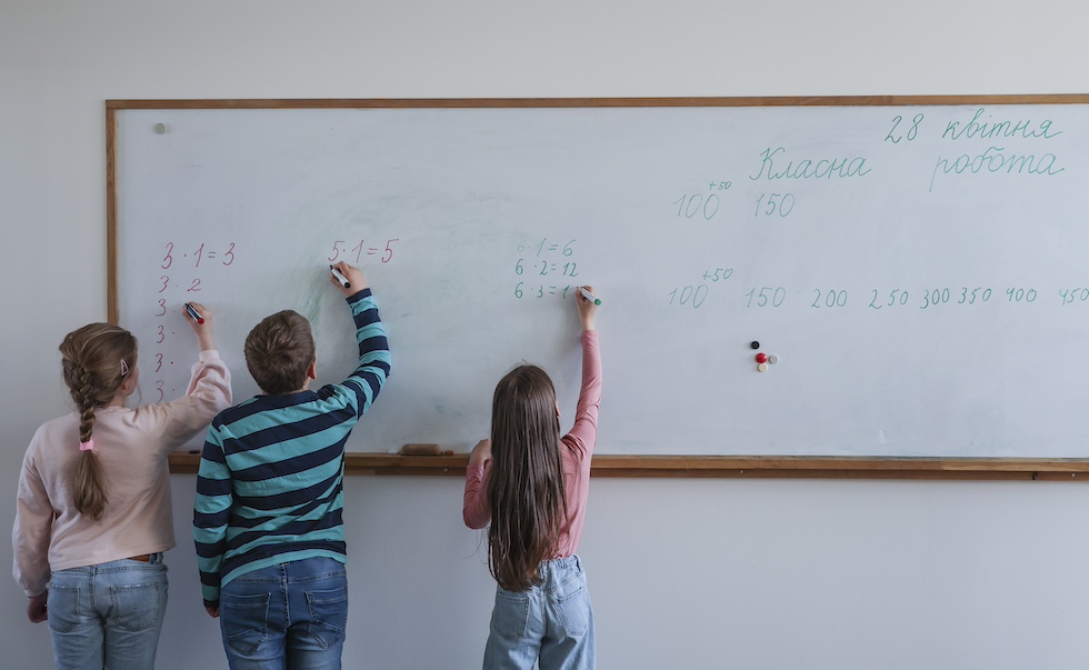 Tre bambini di spalle impegnati a scrivere operazioni aritmetiche alla lavagna