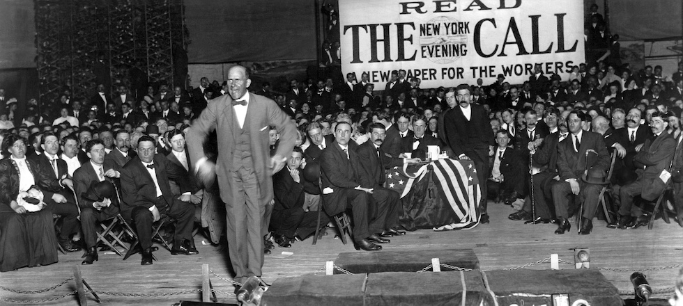 Una foto in bianco e nero in cui Eugene V. Debs fa un discorso in piedi durante una riunione di un Sindacato dei Lavoratori nel 1912. Dietro di lui uno striscione e una platea di uomini seduti che lo ascoltano.