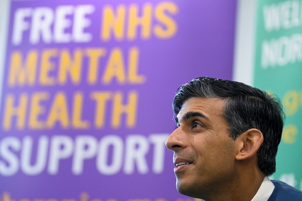 Rishi Sunak in primo piano con un cartellone sullo sfondo che recita "Free NHS Mental Health Support"