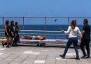 un uomo sdraiato su una panchina di cemento su un marciapiede davanti al mare, di fianco a persone che camminano e si fanno selfie