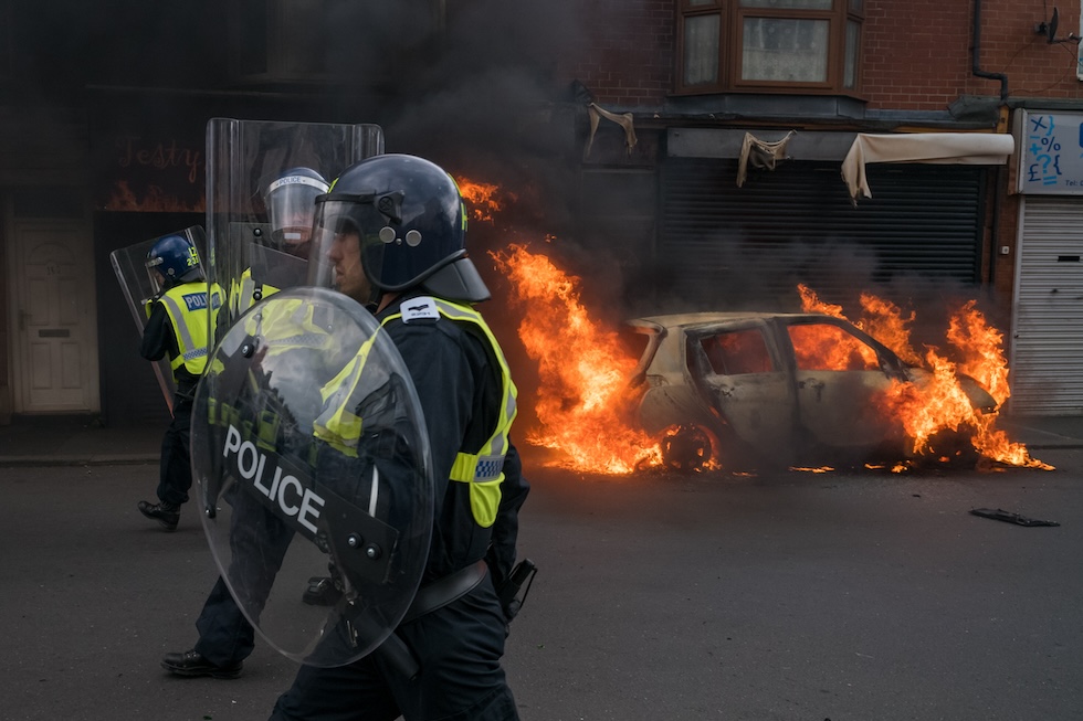 La polizia in assetto antisommossa a fianco di una macchina incendiata durante le proteste a Middlesbrough, il 4 agosto