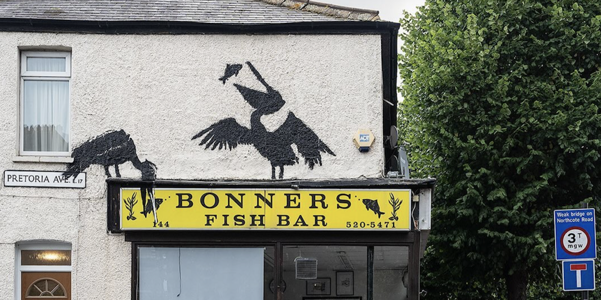 Due sagome nere di pellicani che mangiano dei pesci sopra l'insegna gialla di un negozio di fish and chips chiamato "bonners"