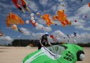 una donna su una spiaggia con un aquilone verde, dietro di lei diversi grossi aquiloni a forma di rana