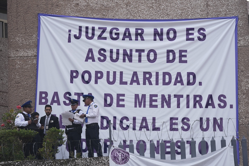 Un grande lenzuolo con uno slogan scritto in stampatello e in viola. In basso a sinistra davanti allo striscione ci sono quattro agenti di sicurezza del tribunale