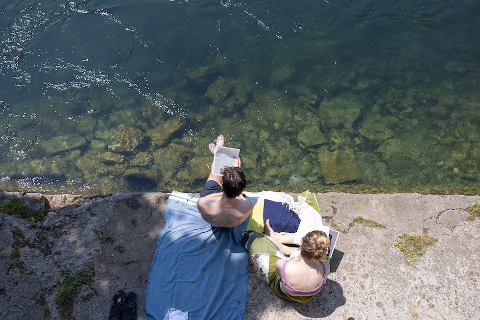 Due persone viste dall'alto leggono vicino al fiume, una di loro con i piedi nell'acqua 