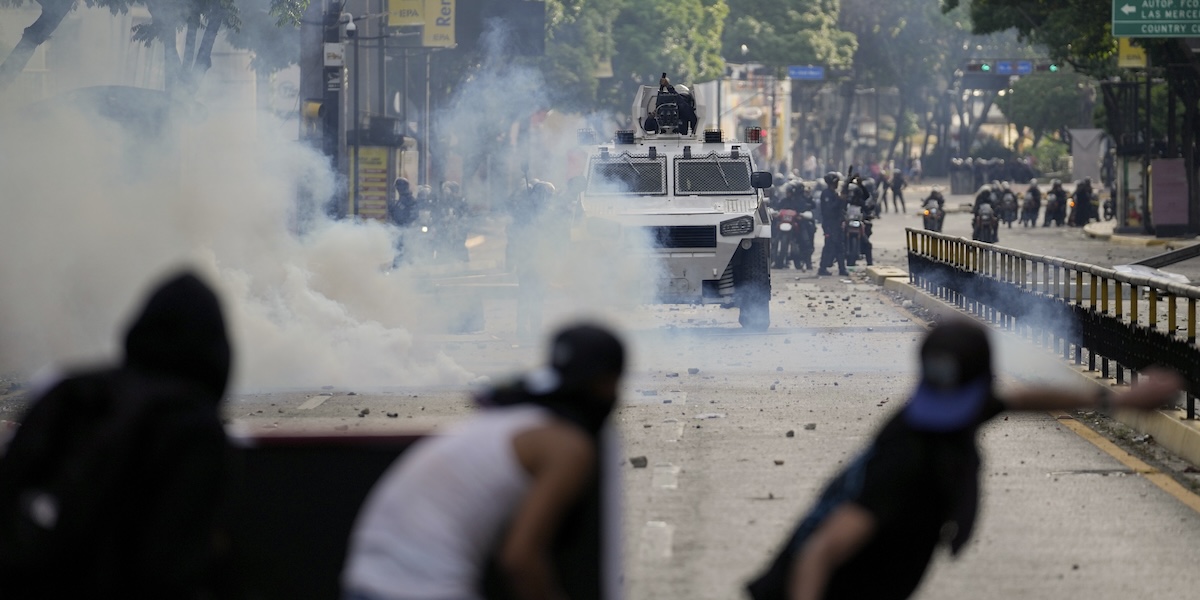 Manifestanti col volto coperto si nascondo dietro una barricata; davanti a loro un mezzo corazzato dell'esercito