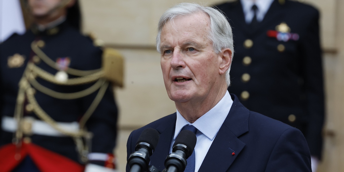 Michel Barnier fotografato a mezzo busto in un completo blu parla al microfono nel cortile dell'Hotel Matignon