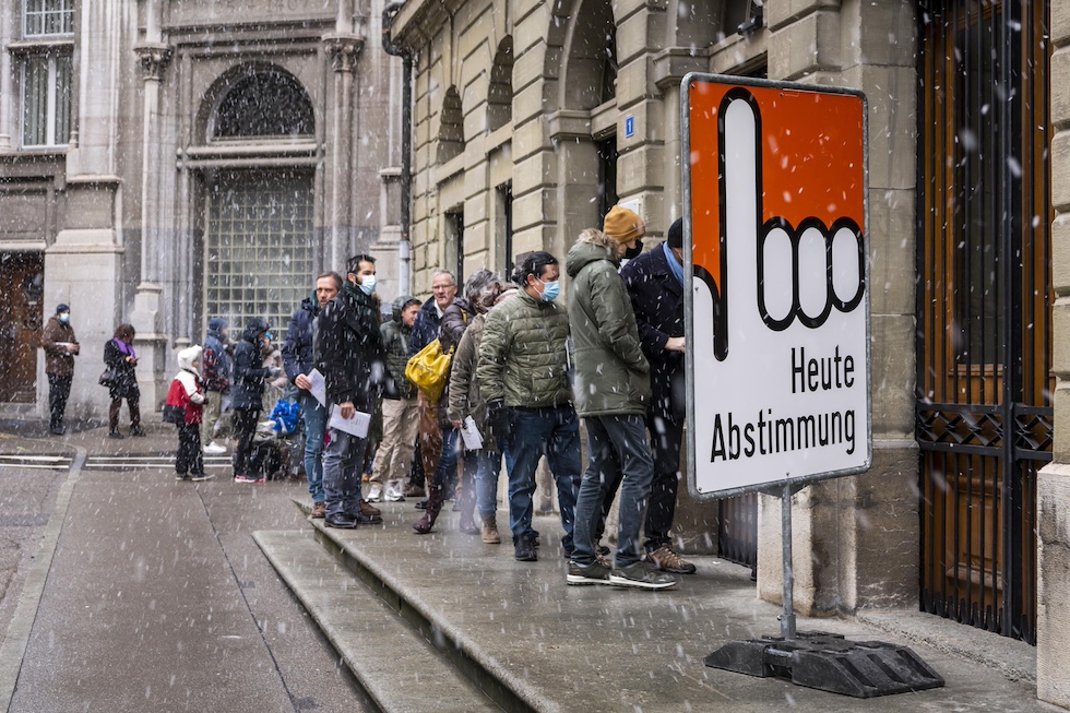 Persone in coda per strada mentre nevica davanti a un cartello con scritto "si vota oggi" in tedesco