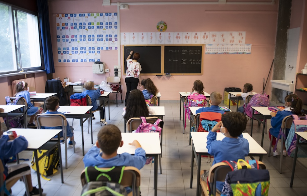 Una classe di bambini e bambine inquadrati di spalle durante la lezione, mentre l'insegnante scrive alla lavagna