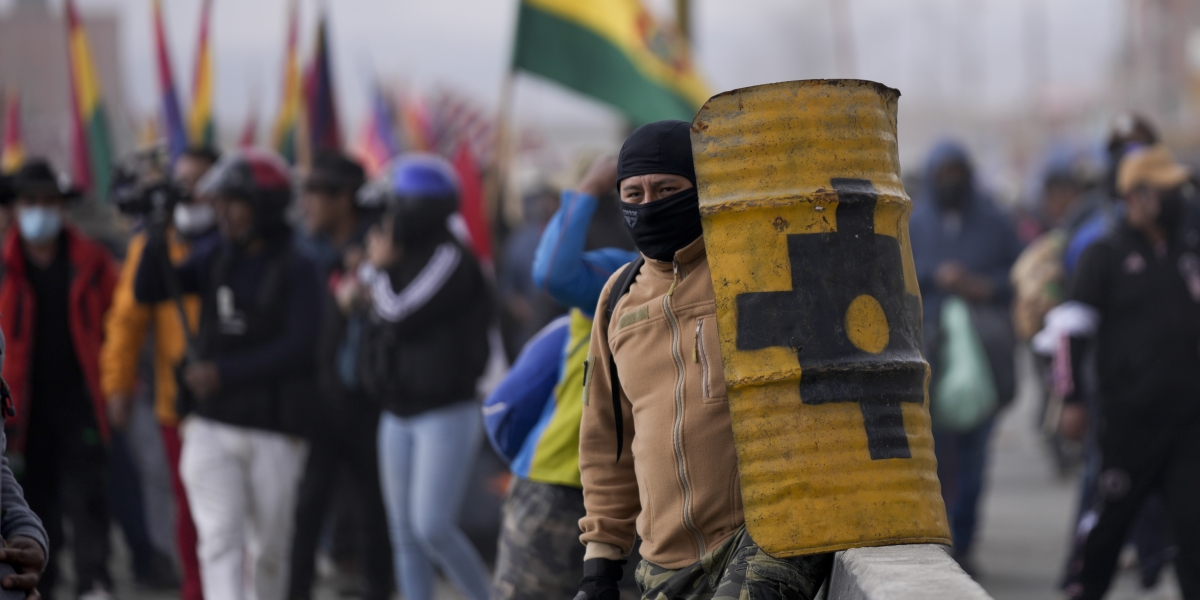 un uomo con un passamontagna e uno scudo antisommossa artigianale fatto con un pezzo di bidone, davanti a una schiera di manifestanti con bandiere