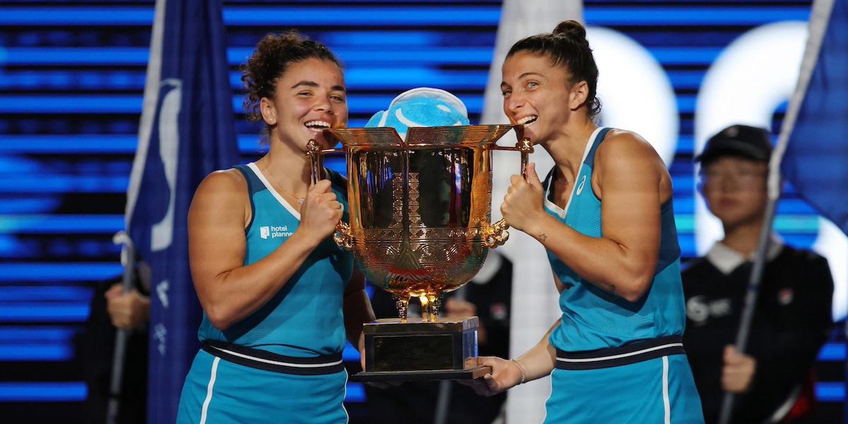 Le due atlete vestite di azzurro stringono una grande coppa d'oro mentre sorridono e la mordono ai bordi, come si usa fare con le medaglie