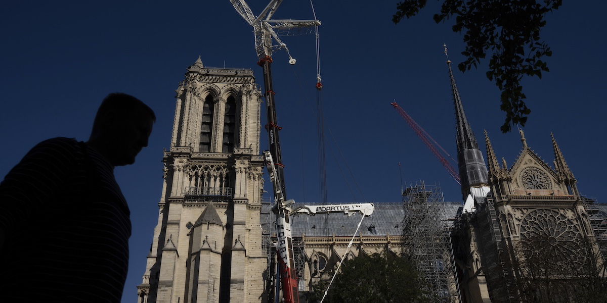 Notre-Dame vista di lato con due gru, sullo sfondo un cielo azzurro scuro. In primo piano in contro luce, sulla sinistra, la figura di un uomo