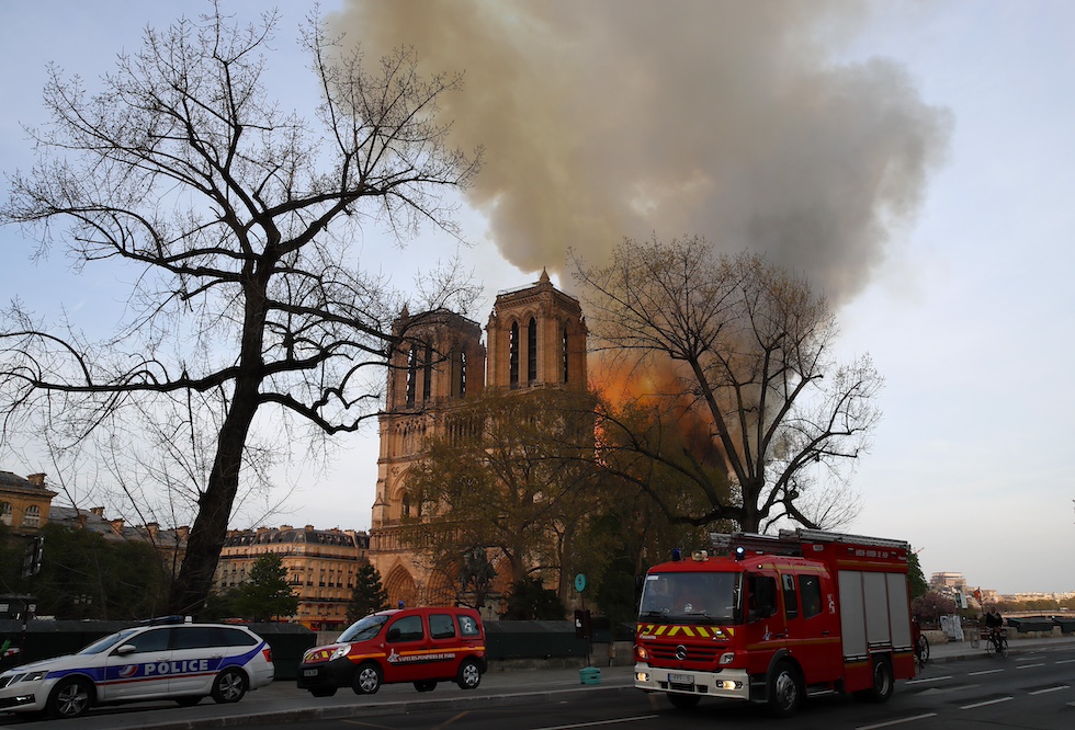 Notre-Dame brucia sullo sfondo, con un grande fumo che si alza in cielo, di giorno. In primo piano ci sono un albero senza foglie e alcune macchine, fra cui un camion dei pompieri