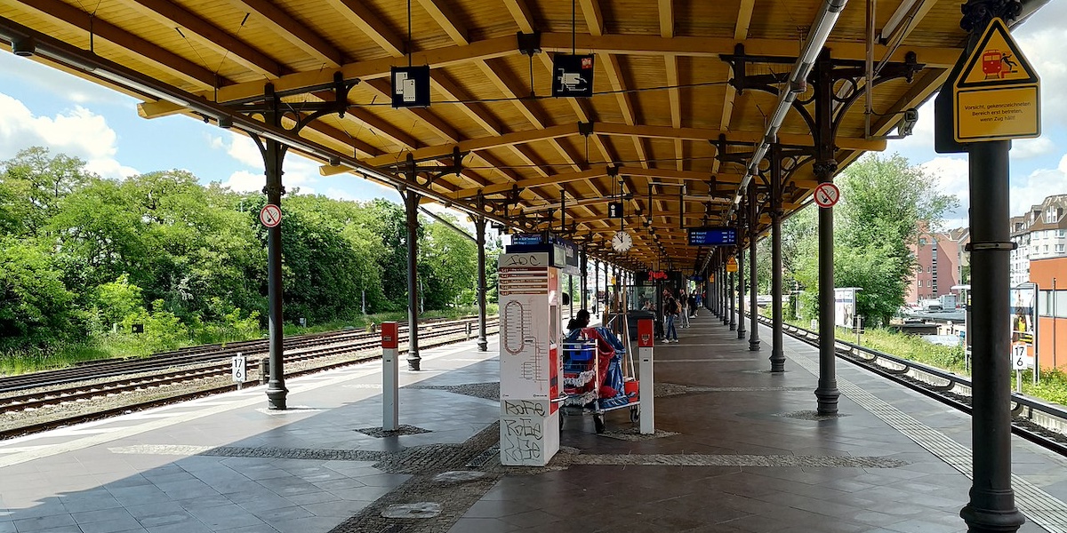 Una banchina di una stazione di treni con un tetto, aperta da entrambi i lati sui binari, circondati da alberi