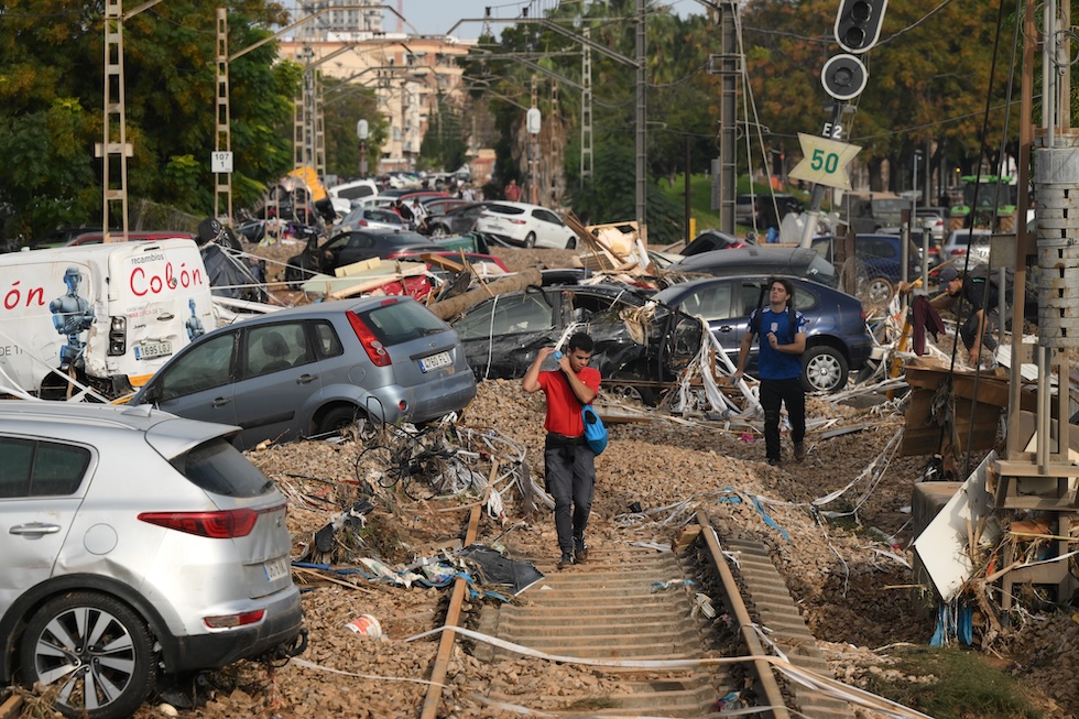 Un uomo trasporta una tanica d'acqua lungo i binari della ferrovia, in buona parte occupati da detriti e automobili trascinate dall'acqua