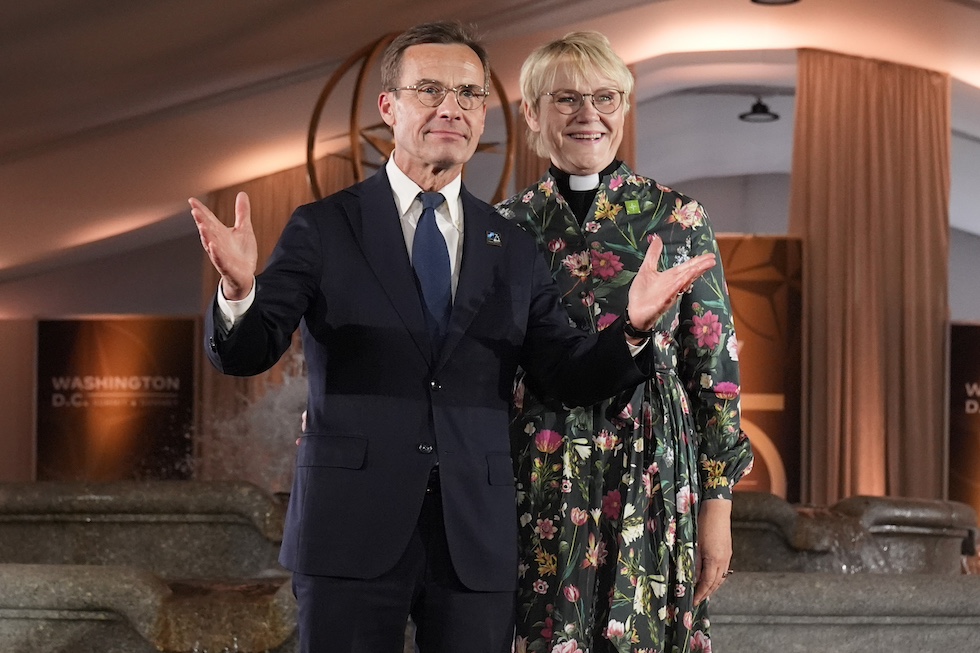 Ulf Kristersson indossa un abito scuro, camicia bianca, cravatta blu e occhiali da vista e ha entrambe le braccia sollevate all'altezza del petto. Brigitta Ed indossa un abito a fiori, il collarino ecclesiastico e degli occhiali da vista. Ha i capelli biondi tagliati corti e sorride.