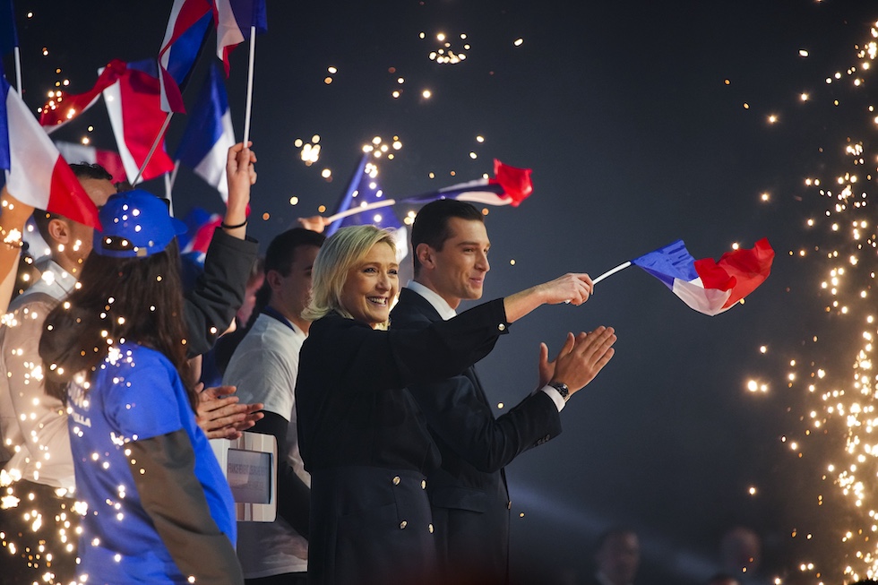 Marine le pen e Jordan bardella di profilo sorridono circondati da bandiere francesi e fuochi d'artificio