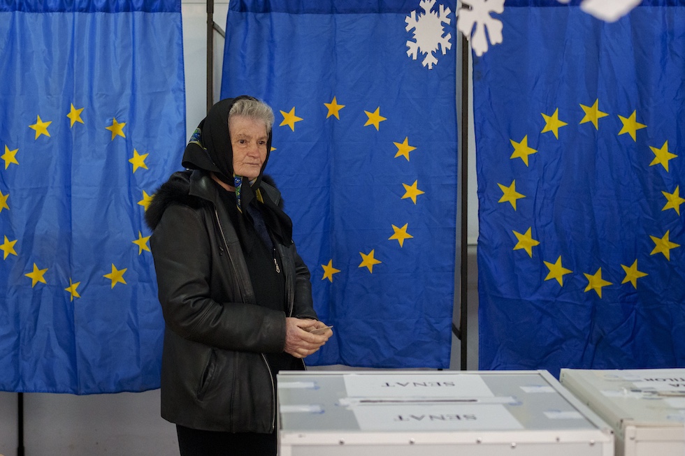 Una donna anziana vestita di nero con un fazzoletto in testa si avvicina a un'urna elettorale dopo aver votato. Dietro di lei le tende dei seggi sono fatte con bandiere dell'Unione Europea