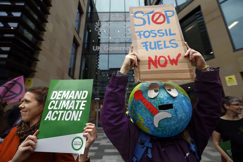 Una manifestante regge un cartello con la scritta “Demand Climate Action” mentre un altro regge un cartello con la scritta “Stop fossil fuels now” e indossa sul volto una maschera del mondo con gli occhi sofferenti e un termometro rosso vicino alla bocca