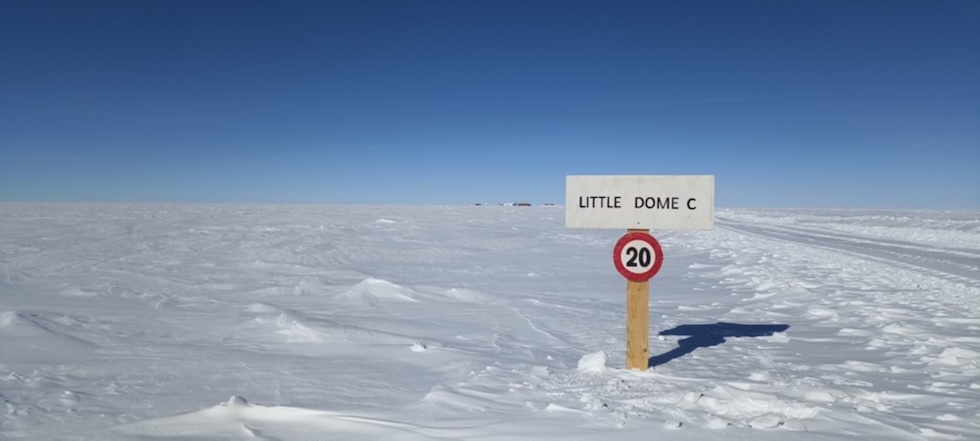 Un cartello con scritto "Little Dome C" e con un divieto di velocità superiori ai 20 chilometri orari in una vasta area pianeggiante; solo all'orizzonte si intravedono delle strutture umane